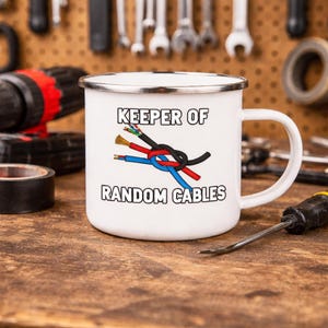 May include: White enamel mug with a black rim and handle. The mug features the text "KEEPER OF RANDOM CABLES" above a graphic of intertwined red, blue, and black electrical wires. The mug is on a wooden surface.
