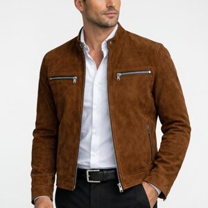 Può includere: Giacca in pelle scamosciata marrone con zip frontale e due tasche con zip sul petto. La giacca è indossata sopra una camicia bianca con colletto e pantaloni neri. È visibile una cintura nera con fibbia argentata.