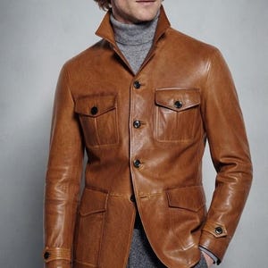 Pode incluir: Uma jaqueta de couro castanho-claro com gola alta, quatro bolsos frontais com aba e botões, e punhos abotoados. A jaqueta é usada sobre uma blusa de gola alta cinza e combinada com calças cinzas, complementada por óculos de sol com lentes castanhas.