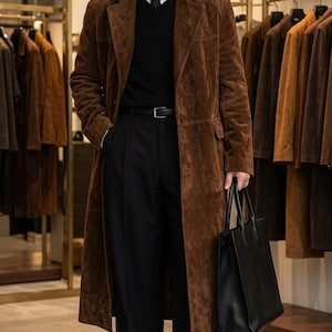 May include: A long brown suede coat worn over a white collared shirt, a black V-neck sweater, and black trousers. The outfit includes a black leather belt, black dress shoes, and a black leather tote bag. The coat features a notched lapel.
