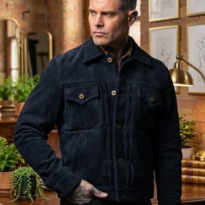 Può includere: Giacca in camoscio blu navy con due tasche sul petto e due tasche laterali. La giacca ha chiusure con bottoni e un colletto classico. La giacca è indossata sopra una camicia marrone.