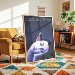 May include: A framed vertical poster featuring a dark blue starry night sky with a crescent moon. The artwork depicts a large, pale, serene face resting on a hand, with a small couple dancing in a pink dress and black suit atop the head. The text reads Verdi La Traviata.