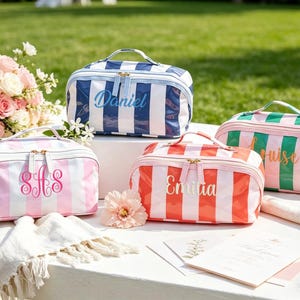Op de afbeelding: Vier gestreepte cosmetische tassen in blauw, roze, rood en groen met witte strepen. Elke tas heeft een handvat en een ritssluiting. De tassen zijn gepersonaliseerd met namen of initialen in een cursief lettertype.
