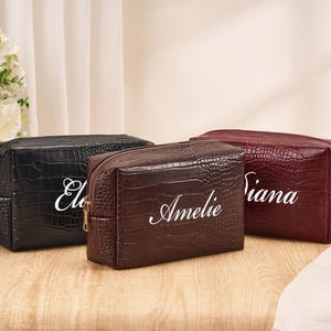 May include: Three rectangular cosmetic bags with a crocodile-embossed texture, featuring gold-tone zippers. The bags are black, dark brown, and burgundy, each personalized with white cursive text reading "Ele", "Amelie", and "Diana" respectively.