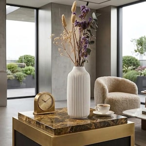 May include: A white, ribbed ceramic vase filled with dried flowers and greenery sits on a marble-topped coffee table. A gold-colored clock and a cup of coffee on a saucer are also on the table. A beige armchair is in the background.