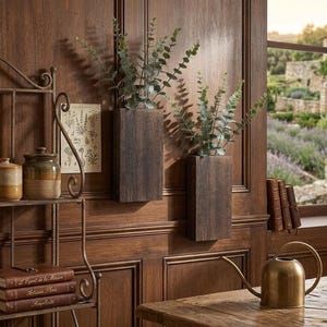 May include: Two dark wooden wall planters, each holding faux greenery, are mounted on a wooden door. The door is a rich, warm brown. A metal shelf with jars and books is on the left. A brass watering can sits on a wooden table.