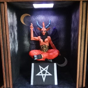 Puede incluir: Escultura de Baphomet, deidad con cabeza de cabra, sentado en una caja con el texto "SOLVE ET COAGULA" arriba. La figura es roja con cuernos azules y alas negras. Un pentagrama está en la base. El fondo es oscuro con acentos de media luna.