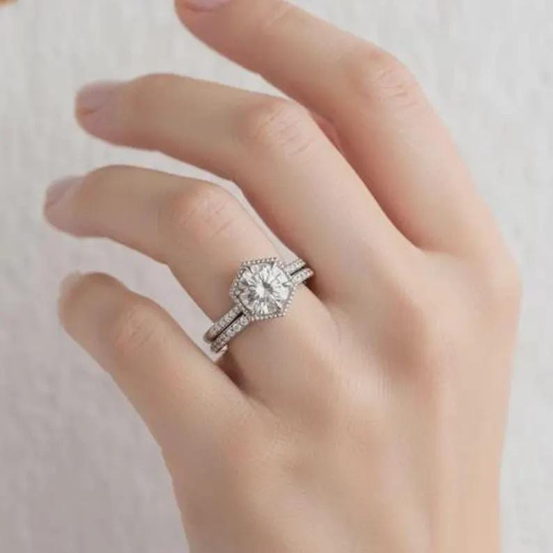 May include: A silver-toned ring set featuring a central hexagonal-shaped setting with a round brilliant-cut stone, surrounded by a halo of small pav&eacute; stones. The band is also adorned with a row of small pav&eacute; stones along the shank.