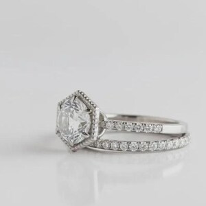 May include: A silver-toned ring featuring a central round gemstone set within a hexagonal frame. The band is split into two distinct sections, with the lower band and the upper section of the hexagonal setting adorned with small, clear, round stones.
