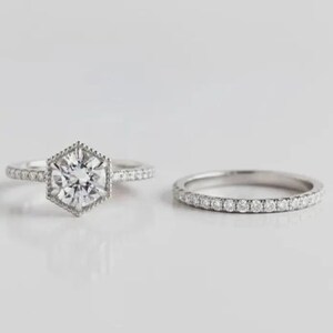 May include: A silver-toned engagement ring set featuring a central round brilliant-cut stone held in a hexagonal setting with milgrain detailing, accompanied by a matching pav&eacute;-set band. Both rings feature small clear stones along the bands.