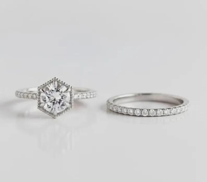 May include: A silver-toned engagement ring set featuring a central round brilliant-cut stone held in a hexagonal setting with milgrain detailing, accompanied by a matching pav&eacute;-set band. Both rings feature small clear stones along the bands.