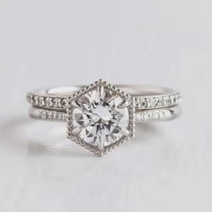 May include: A silver-toned ring set featuring a central round-cut clear stone held in a hexagonal, beaded-edge setting. The band and the accompanying stackable ring are both adorned with small, clear pav&eacute;-set stones along the outer surface.