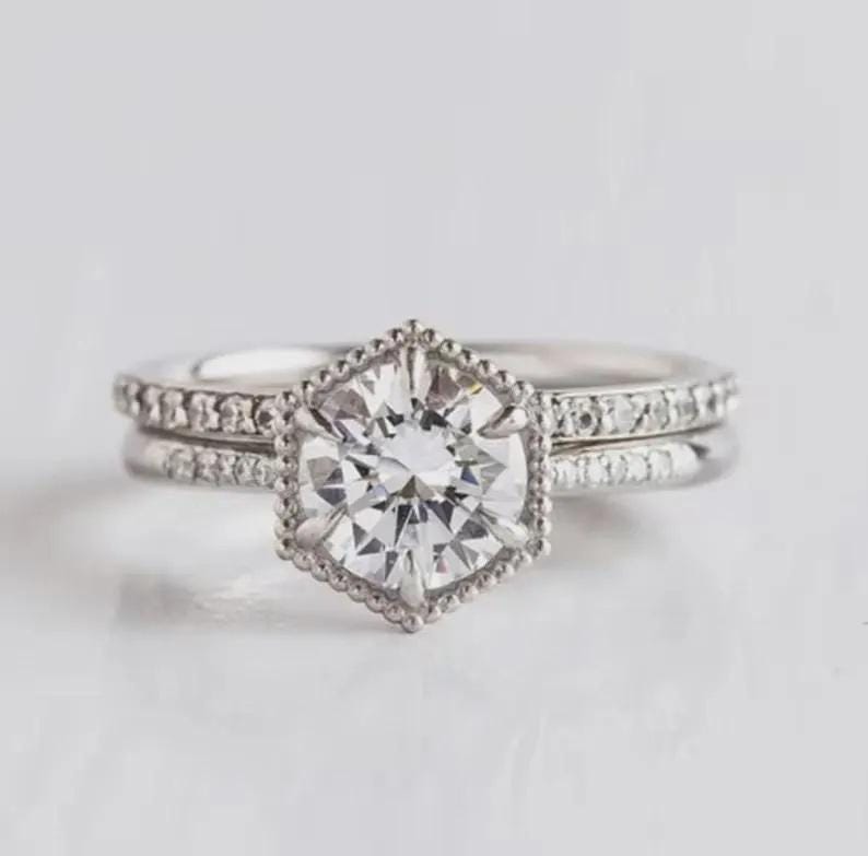 May include: A silver-toned ring set featuring a central round-cut clear stone held in a hexagonal, beaded-edge setting. The band and the accompanying stackable ring are both adorned with small, clear pav&eacute;-set stones along the outer surface.