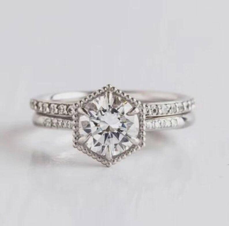 May include: A silver-toned ring featuring a central round-cut stone set within a hexagonal, beaded frame. The band is adorned with a row of small, sparkling stones along the shank, creating a detailed and reflective appearance on the metal surface.
