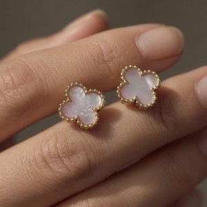 May include: A pair of clover-shaped stud earrings. Each earring has a light pink, pearlescent centre framed by a gold beaded border. The earrings are shown against a neutral background.