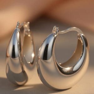 May include: A pair of polished silver hoop earrings. The earrings have a smooth, teardrop shape and a shiny, reflective surface. The earrings are designed with a hinged closure.