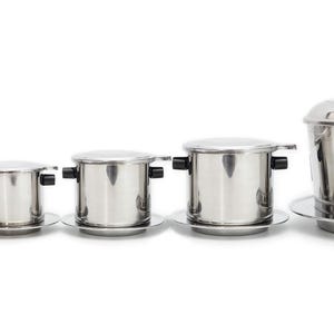 May include: A set of five stainless steel Vietnamese coffee filters arranged in a row from smallest to largest. Each cylindrical coffee dripper features a matching lid with a small black knob, a perforated base plate, and two small black side handles.