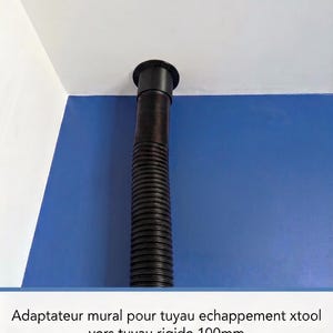 Può includere: Un adattatore a parete nero per tubo di scarico xTool verso un tubo rigido da 100mm. L'adattatore è montato a filo contro un soffitto bianco, con un tubo flessibile corrugato nero che si estende verso il basso su una parete blu.