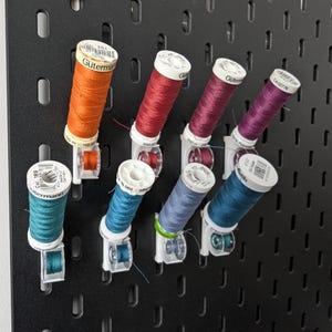 May include: A black pegboard displaying spools of colorful thread. The thread colors include orange, red, purple, teal, blue, and teal. The spools are labeled with the brand name "Gütermann".