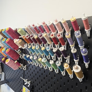 Puede incluir: Un panel perforado negro de pared con numerosos ganchos de plástico blanco que sostienen una variedad de carretes de hilo de coser de colores. Los carretes están dispuestos en filas y columnas, mostrando un espectro de colores como rosa, azul, amarillo y morado.
