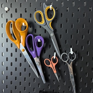 May include: A collection of scissors in various sizes and colors, including orange, purple, and gray, hanging on a black pegboard. The scissors have different handle designs and blade lengths, suitable for various cutting tasks.