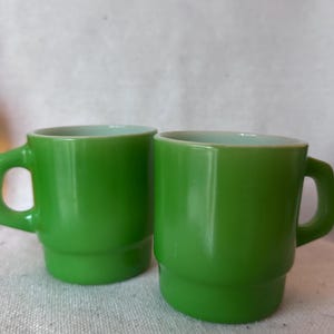 May include: Two bright lime green glass mugs with a smooth, glossy finish. Each mug features a cylindrical body with a slightly wider base, a rounded handle on the side, and a clean, simple design. The glass has a semi-translucent quality.