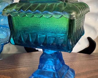 Puede incluir: Un dulcero cuadrado de vidrio con tapa, que presenta un degradado vibrante desde el azul profundo en la base hasta el verde brillante en la parte superior. La pieza tiene una superficie texturizada, un borde festoneado y una base cuadrada.