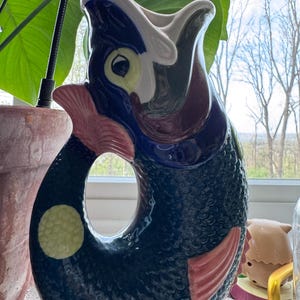 May include: A ceramic pitcher in the shape of a fish, featuring a dark blue body with textured scales. The fish has red fins, a yellow spot, and a white and blue open mouth. The pitcher is a decorative item.