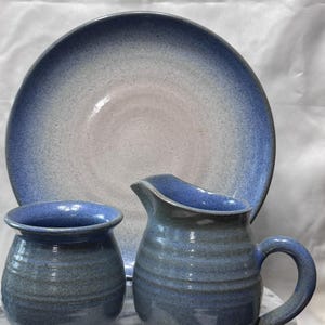 May include: A ceramic set consisting of a large round plate, a small rounded mug, and a pitcher with a handle. Each piece features a speckled glaze with a gradient transition from a deep blue rim to a lighter, pale center on the plate.