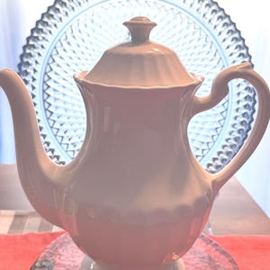May include: A white porcelain teapot featuring a fluted design on the lid and base, a curved handle, and a long, elegant spout. The teapot has a smooth, glossy finish and a classic silhouette, standing on a circular pedestal base with a matching lid.