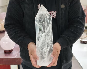 Può includere: Una grande punta di cristallo di quarzo trasparente, tenuta da una persona. Il cristallo è trasparente con inclusioni interne e una parte superiore appuntita. Altre sfere di cristallo sono visibili sullo sfondo.