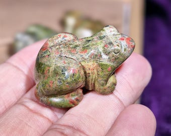 Può includere: Una statuetta a forma di rana in pietra scolpita nei toni del verde, rosso e beige. La rana è in posizione seduta con dettagli precisi. La statuetta è tenuta in una mano, con uno sfondo sfocato.