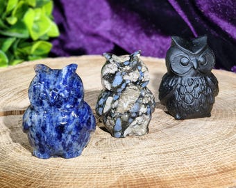 Può includere: Tre statuette di gufo scolpite nella pietra. La prima è blu scuro con venature bianche, la seconda è un mix di grigio, bianco e blu, e la terza è nera. Sono esposte su una superficie di legno.