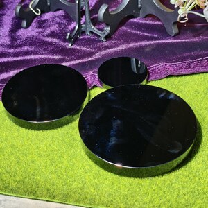 May include: Three black, circular display stands of varying sizes. The stands have a reflective, glossy surface and a silver-colored rim. They are arranged on a green surface, with a purple velvet cloth and a decorative stand in the background.