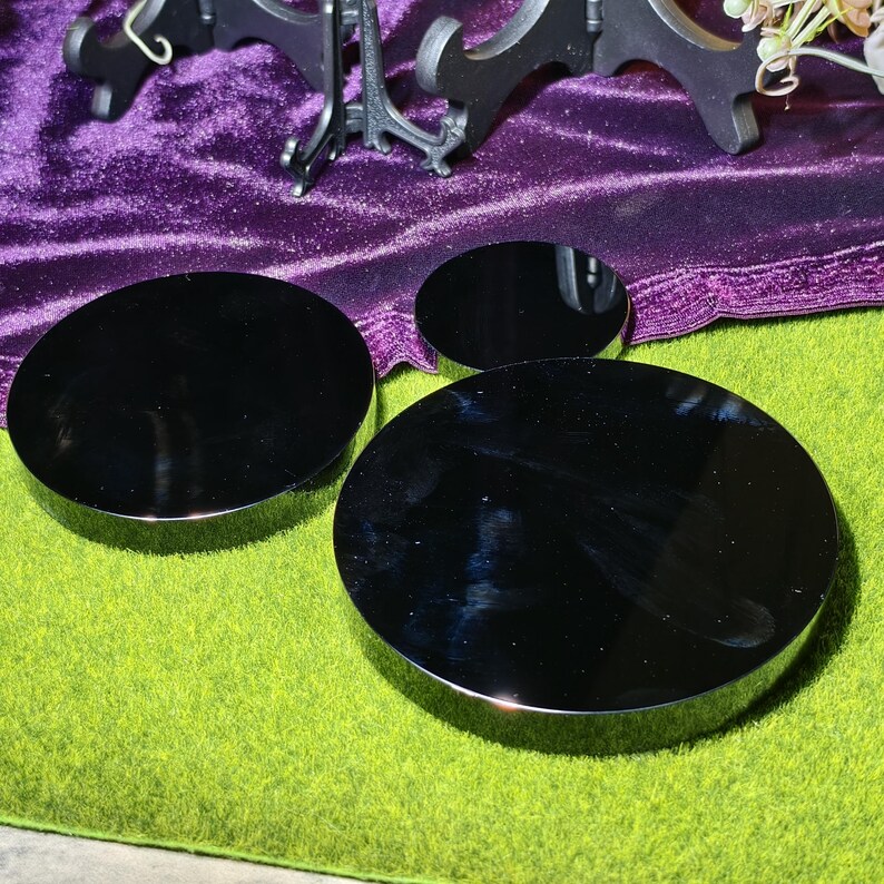 May include: Three black, circular display stands of varying sizes. The stands have a reflective, glossy surface and a silver-colored rim. They are arranged on a green surface, with a purple velvet cloth and a decorative stand in the background.