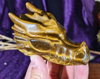 Può includere: Una scultura in pietra preziosa occhio di tigre intagliata a forma di testa di drago. La pietra presenta un colore marrone dorato con una lucentezza setosa e chatoyante e distinte striature marrone scuro. L'intaglio mostra squame, corna e muso.