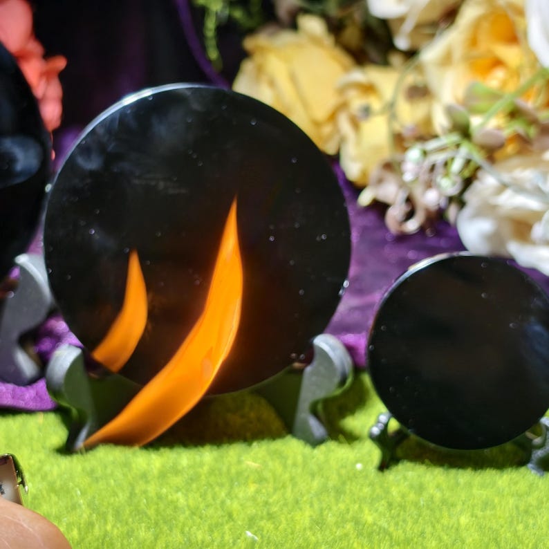 May include: Black, round decorative objects with orange flame-like designs, displayed on small stands. The objects are set against a backdrop of flowers and a green surface. The overall theme suggests a decorative or artistic product.