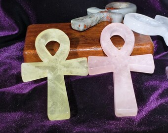 May include: An assortment of ankh-shaped stone carvings. The ankhs are in various colors, including pale yellow and pink. They are displayed on a wooden block and a purple velvet surface.