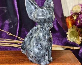 Può includere: Una statuetta in pietra levigata scolpita a forma di gatto seduto con grandi orecchie a punta e una superficie liscia. La pietra presenta un motivo maculato di grigio scuro, nero e bianco su tutto il corpo e la testa della statuetta.
