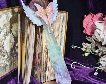 May include: A decorative bookmark featuring a pink butterfly with white wings, a heart-shaped accent, and a large, multi-colored crystal. The bookmark is placed in an open book with floral illustrations, set against a dark purple background.