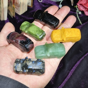 May include: A collection of small, carved stone car figurines in various colors, including black, green, yellow, and a speckled blue and gray. The miniature cars are held in a hand, showcasing their detailed craftsmanship and polished surfaces.