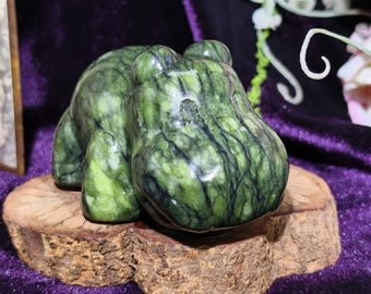 Può includere: Una statuetta di ippopotamo in pietra verde scolpita con venature scure, esposta su una piccola base di legno rotonda. La scultura ha una finitura lucida ed è ambientata su uno sfondo di velluto viola. Un oggetto decorativo unico.