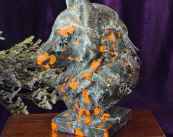 Può includere: Una scultura in pietra scolpita raffigurante la testa di un lupo. La scultura è prevalentemente grigia con macchie arancioni e nere vivaci. È montata su una base quadrata e poggia su una superficie di legno.