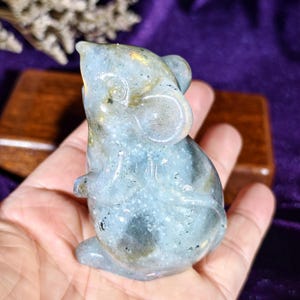 Pode incluir: Uma estatueta de rato de labradorite esculpida. O rato é azul claro com manchas douradas e azul escuro. A estatueta é segurada em uma mão, com um fundo roxo e um bloco de madeira visível atrás do rato.