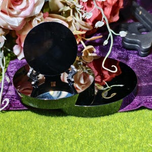 May include: A reflective, black, circular object is supported by a silver-colored, decorative stand. The stand sits on a reflective, silver-colored, semi-circular base. The background includes flowers and a purple velvet cloth.