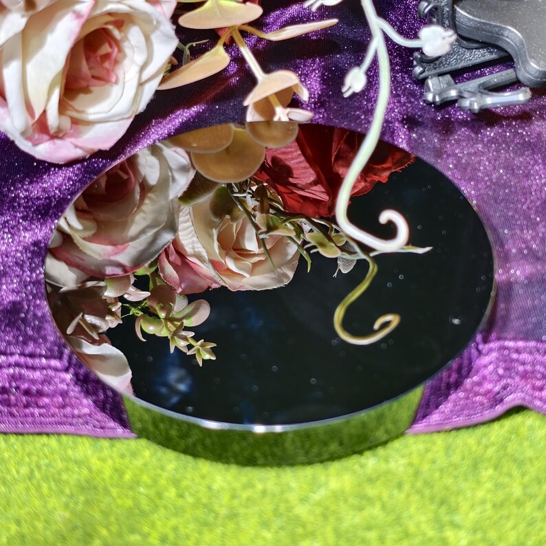 May include: A round, silver-rimmed mirror reflecting a bouquet of pink and red roses and other flowers. The mirror sits on a bright green surface, with a purple, glittery fabric in the background.