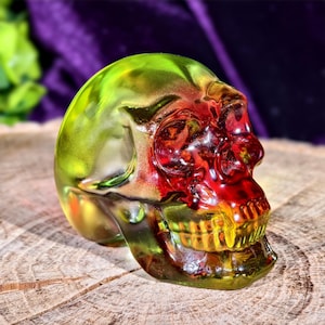 May include: A decorative glass skull with a gradient of colors, including green, yellow, and red. The skull is resting on a wooden surface. The skull's details are visible, including the eye sockets, nose cavity, and teeth.