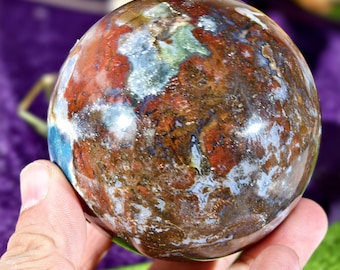 Può includere: Una pietra sferica lucida caratterizzata da un motivo complesso di inclusioni rosso scuro, marrone terra, blu tenue e bianco crema. La superficie è liscia e riflettente, mostrando una consistenza marmorizzata con vari depositi minerali.