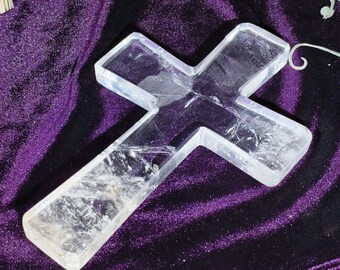 Può includere: Una croce di cristallo di quarzo trasparente con fratture interne visibili. La croce è posta su uno sfondo di velluto viola scuro. Il cristallo è trasparente, lasciando passare la luce, e ha una superficie liscia e lucida.