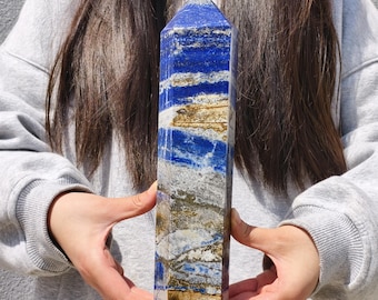 Può includere: Un obelisco rettangolare di cristallo di lapislazzuli. La pietra lucida è di un blu intenso con striature bianche, dorate e grigie. L'obelisco è tenuto in posizione verticale, mostrando la sua forma geometrica e i suoi motivi naturali. Circa 20 cm di altezza.
